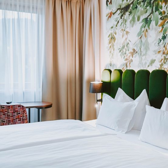 Ein Hotelzimmer mit einem Doppelbett, das mit weißen Laken und Kissen bedeckt ist. Eine Lampe steht auf dem Nachttisch. In der Nähe des Fensters befindet sich ein Schreibtisch mit einem Stuhl.