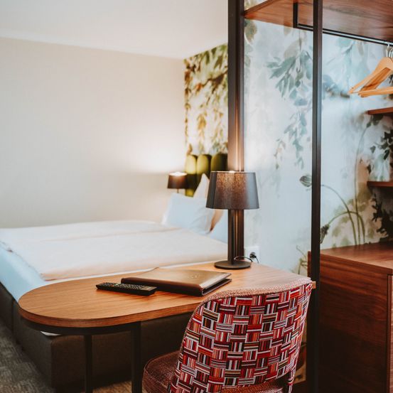 Ein Schlafzimmer mit einem Bett, einem Schreibtisch mit Fernbedienung und einem Stuhl. Die Wand hat ein Tapetendesign. Eine Lampe und zwei Kissen sind auf dem Bett.