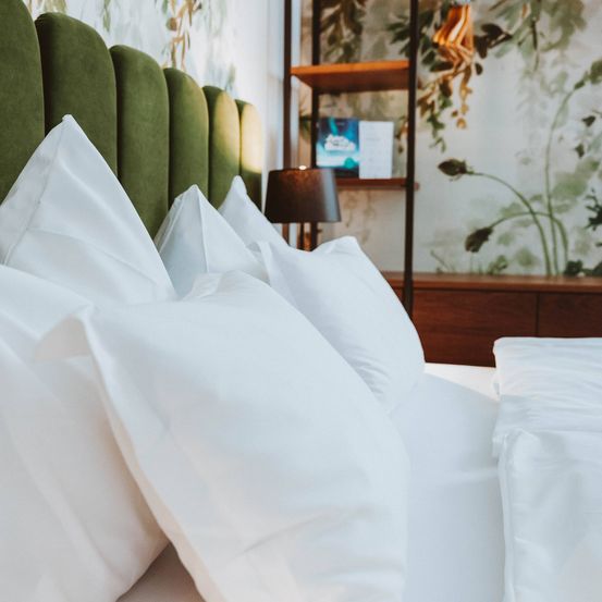 Ein Bett mit weißen Kissen und einem grünen Kopfteil ist ordentlich gemacht. Eine Lampe steht auf dem Nachttisch, und ein hölzernes Regal hält ein Buch. Die Wand ist mit einem botanischen Tapetenmuster verziert.