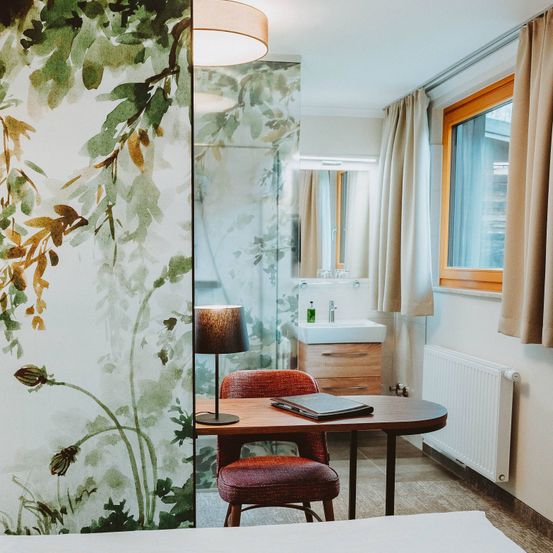 Ein Innenraum eines Hotelzimmers mit Schreibtisch, Stuhl, Lampe und gemusterter Tapete. Das Zimmer verfügt auch über Waschbecken, Spiegel und Fenster.