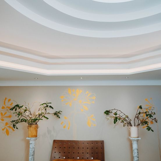 Ein Raum mit weißen Wänden, einem gewölbten Deckenbogen und gelben Blumendesigns. Zwei Topfpflanzen auf Marmorsockeln flankieren einen braunen Stuhl.