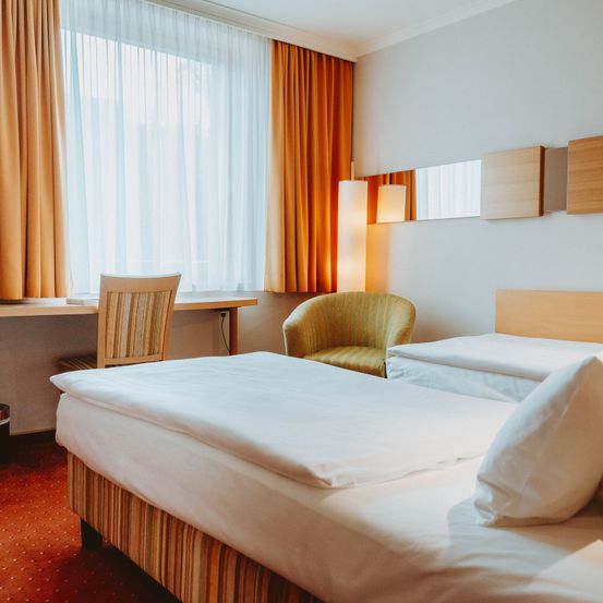 Ein Hotelzimmer verfügt über zwei Betten, einen Schreibtisch mit Stuhl und einen Stuhl am Fenster. Der Raum ist gut beleuchtet mit weißen Vorhängen und Holzpaneelen an der Wand.