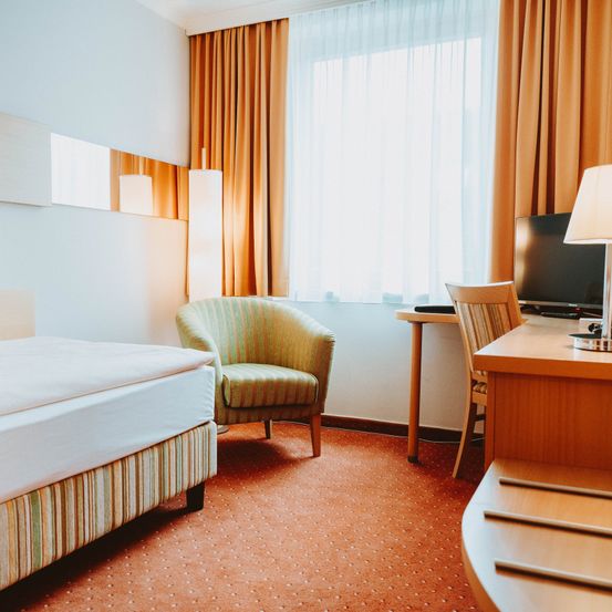 Ein Hotelzimmer verfügt über ein Bett mit weißen Laken, einen gestreiften Stuhl, einen Schreibtisch mit Monitor und eine Lampe. Das Zimmer hat weiße Wände, einen orangefarbenen Teppich und Vorhänge.
