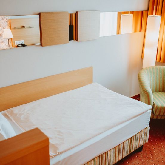 Ein Hotelzimmer mit einem Einzelbett, einem Holzkopfteil, weißen Laken und einem grün gestreiften Sessel. Das Zimmer hat eine weiße Wand, Spiegel und einen Stehlampen.