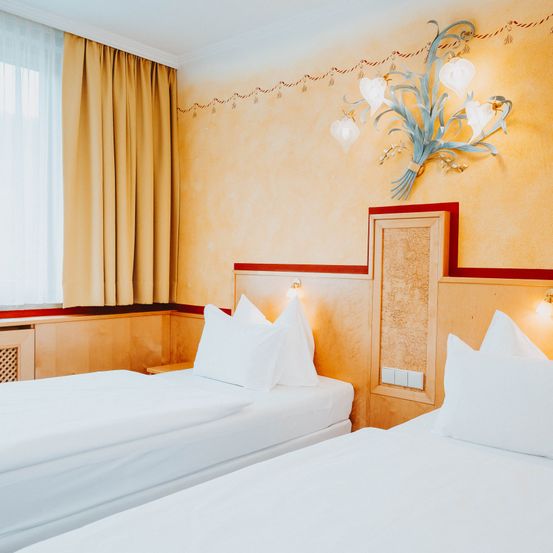 Zwei Einzelbetten mit weißer Bettwäsche und Kissen befinden sich in einem Zimmer mit gelber Wand und weißem Vorhang. Über den Betten befindet sich eine dekorative Wandlampe.