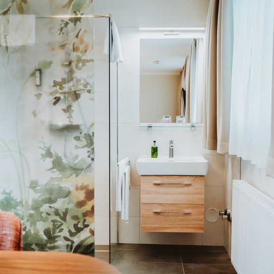 Ein modernes Badezimmer mit einer Duschkabine mit Blattmuster und einem weißen Waschbecken mit Wasserhahn. Ein Spiegel und ein weißes Handtuch sind an der Wand montiert.