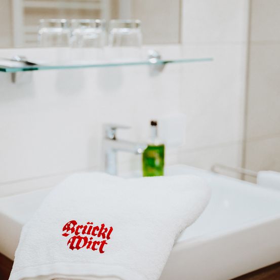 Ein Badezimmer-Waschbecken mit einem weißen Handtuch, auf dem in roten Buchstaben 'Scrunkly Dirt' steht. Das Becken ist sauber, und darüber befindet sich ein Glasregal mit drei Gläsern. Ein Wasserhahn mit einer Flasche grüner Flüssigkeit ist sichtbar.