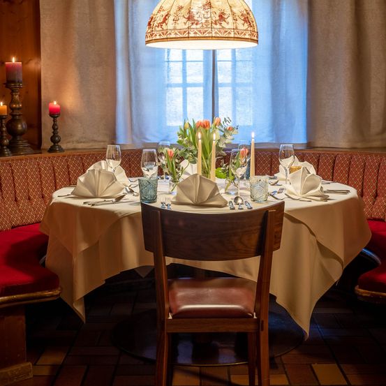 Ein gemütlicher Essbereich mit einem Tisch für zwei Personen, geschmückt mit weißem Tuch, Kerzen und Weingläsern. Die Umgebung umfasst eine Bank mit roten Kissen und einer Lampe.