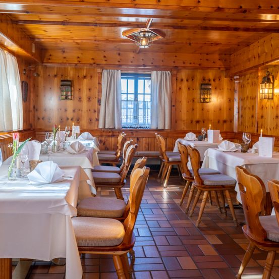 Ein leeres Restaurant mit Holzwänden und für Gäste gedeckten Tischen. Jeder Tisch hat eine weiße Tischdecke, eine Kerze und ein Blumenarrangement. Der Raum wird von einer hängenden Leuchte beleuchtet.