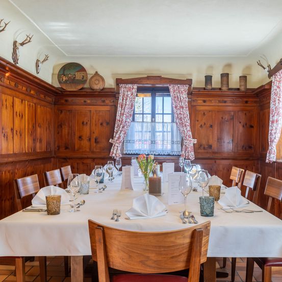 Ein Esszimmer mit Holzwänden und einem Tisch, der für eine Mahlzeit mit Weingläsern, Tellern und Besteck vorbereitet ist. Ein Fenster mit Vorhängen und eine Blumenvase auf dem Tisch.