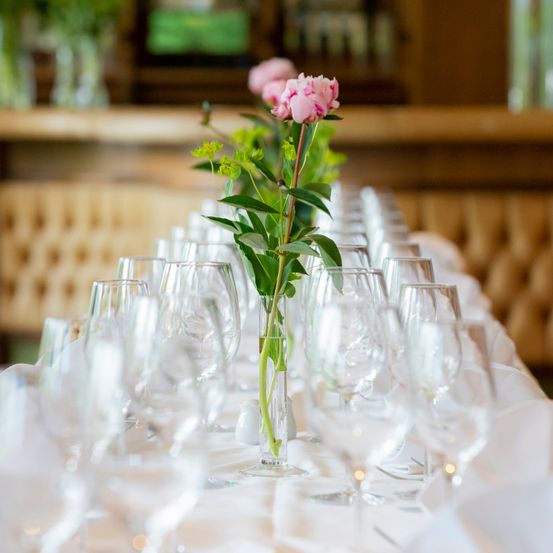 Ein langer Tisch, gesäumt von vielen akkurat aufgestellten Weingläsern, mit einer Vase voller rosa Blumen in der Mitte. Der Tisch ist mit weißem Tuch bedeckt. Dahinter zeigt der verwaschene Hintergrund einen hölzernen Bartresen und ein Fenster.