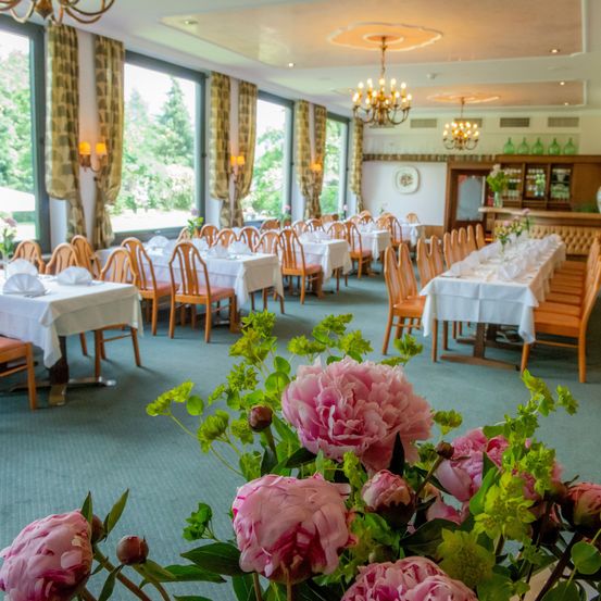 Ein Restaurant mit Reihen von Tischen und Stühlen, geschmückt mit weißen Tischdecken, Blumen und Kronleuchtern. Der Raum verfügt über Glasfenster mit Vorhängen und eine Holzbar.