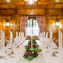 Ein langer Esstisch für eine Mahlzeit mit Blumen und Weingläsern. Holzwände und ein Fenster mit Vorhängen.