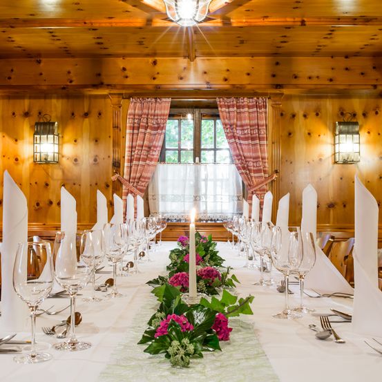 Ein langer Esstisch für eine Mahlzeit mit Blumen und Weingläsern. Holzwände und ein Fenster mit Vorhängen.