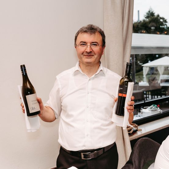 Ein Mann in einem weißen Hemd und mit Brille hält zwei Weinflaschen in den Händen.