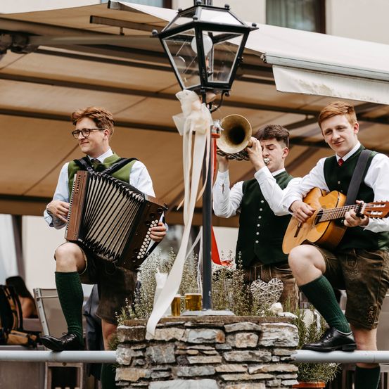 Drei Männer in traditioneller deutscher Kleidung spielen auf einem Balkon, einer spielt Akkordeon, ein anderer Trompete und der dritte Gitarre. Sie sind von dekorativen Elementen umgeben.