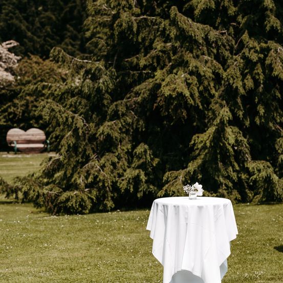 Ein abgedeckter Tisch mit weißer Tischdecke steht auf einem grünen Rasen. Ein Blumenvase steht auf dem Tisch und eine herzförmige Bank ist in der Ferne.