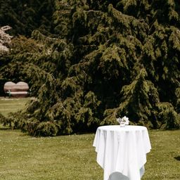Ein abgedeckter Tisch mit weißer Tischdecke steht auf einem grünen Rasen. Ein Blumenvase steht auf dem Tisch und eine herzförmige Bank ist in der Ferne.