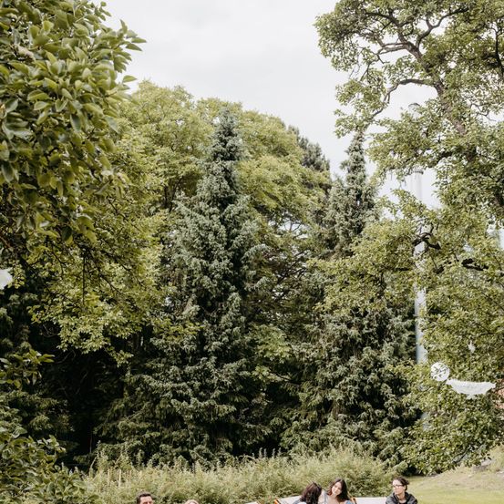 Vier Personen sitzen auf einer Parkbank in einem Park mit grünen Bäumen und Pflanzen im Hintergrund. Das Wetter ist bewölkt.