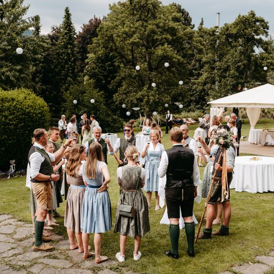 Eine Gruppe von Menschen in traditioneller bayerischer Kleidung versammelt sich in einem Garten für eine Feier, hält Gläser mit Wein und einen Blumenstrauß. Ein weißes Zelt ist aufgestellt, und die Umgebung ist mit Grünpflanzen geschmückt.