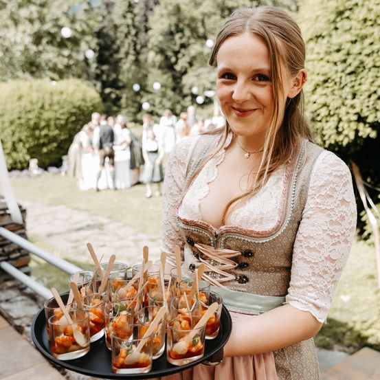 Eine Frau in traditioneller deutscher Kleidung, die einen Tablett mit kleinen Speisen und Holzlöffeln hält, lächelt in die Kamera. Dahinter ist eine verschwommene Outdoor-Veranstaltung mit Menschen zu sehen.