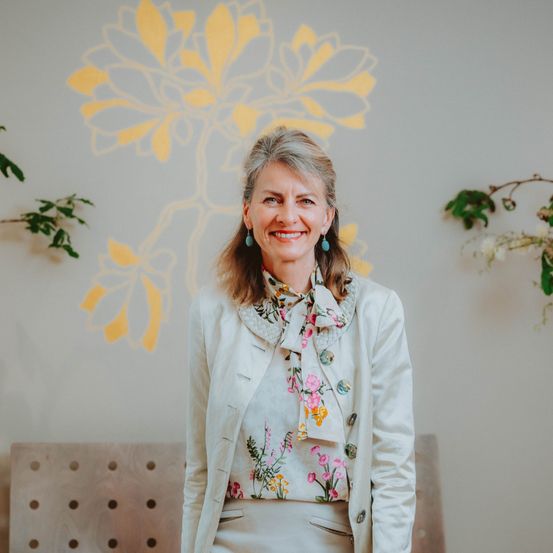 Eine Frau in einer weißen Jacke und einem Blumentopf steht vor einer Wand mit einem gelben Blumendesign.