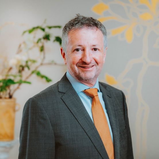 Ein Mann in formeller Kleidung mit einer orangefarbenen Krawatte steht lächelnd in einem Büro mit einem gelben Blumenmuster an der Wand und einer Pflanze im Hintergrund.