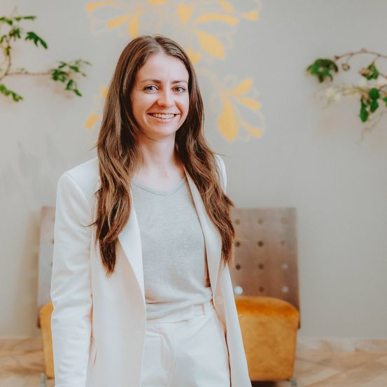 Eine Frau mit langen braunen Haaren lächelt und steht vor einer weißen Wand mit gelben Blumenmustern. Sie trägt einen weißen Blazer und Hosen. Hinter ihr steht ein brauner Stuhl mit einer weißen Sitzfläche.