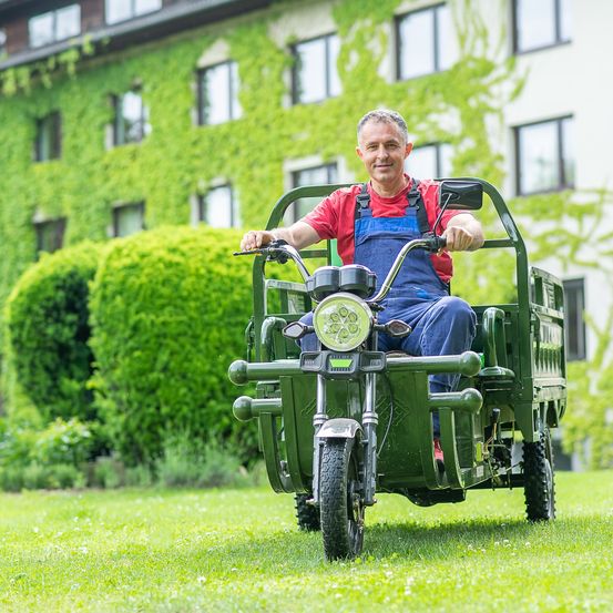 Ein Mann in blauer Latzhose und einem roten Hemd fährt ein grünes dreirädriges Fahrzeug auf einem Rasen vor einem großen Gebäude mit grünem Efeu.