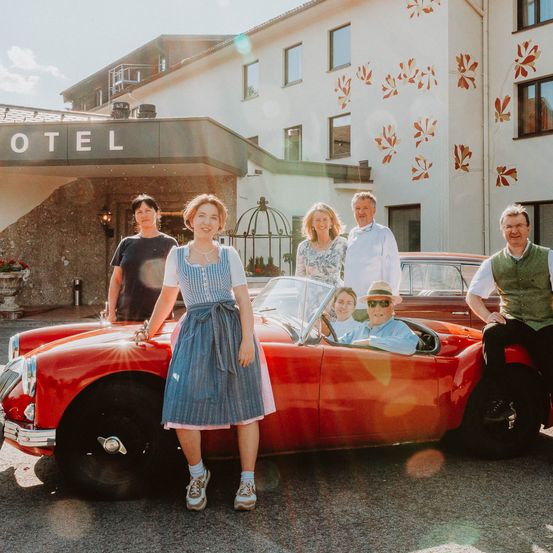 Eine Familie posiert vor einem Hotel. Die Familie besteht aus zwei Frauen, zwei Männern und einem älteren Mann in einem Cabrio. Sie alle lächeln.