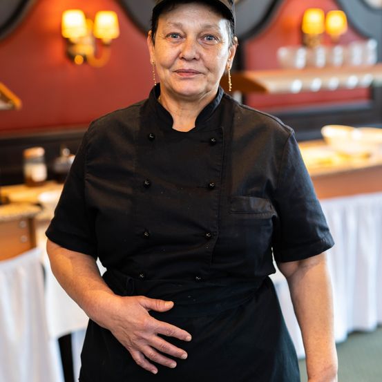 Eine ältere Frau in einem Kochuniform steht in einer Restaurantküche und lächelt in die Kamera. Hinter ihr ist ein Tisch mit weißen Tüchern und verschiedenen Küchenutensilien zu sehen. Die Wand ist rot gestrichen und mit Lampen verziert.
