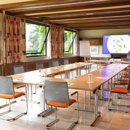 Ein Konferenzraum mit Holzboden, großen Fenstern und Vorhängen. Es gibt lange Tische mit Stühlen, einen Bildschirm an der Wand und eine Tafel.