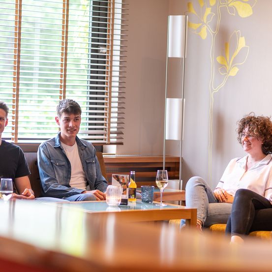 Drei Personen sitzen auf einer Couch und Stühlen und lächeln. Ein Tisch davor hat Weingläser, eine Flasche und eine Kerze. Dahinter ein Fenster mit Jalousien und ein Blumenwanddesign.