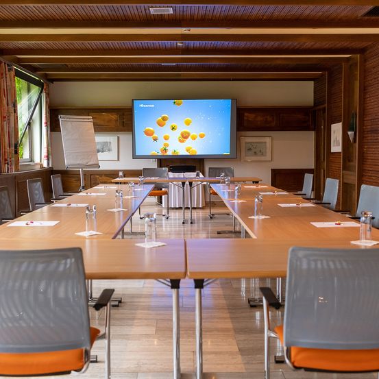 Ein Konferenzraum mit einem großen Bildschirm an der Wand, langen Tischen und Stühlen. Links steht eine Tafel und an der Wand hängen gerahmte Bilder.