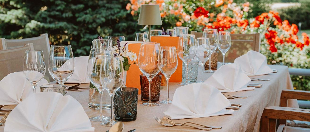 Ein Tisch mit Weingläsern, Servietten und Besteck. Orange Servietten und Gläser in der Mitte. Es gibt eine Lampe und Blumen auf dem Tisch. Bäume und Blumen im Hintergrund.