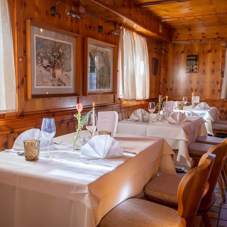 Ein gemütlicher Essbereich mit Holzwänden und -böden. Die Tische sind mit weißen Tischdecken, gefalteten Servietten und Weingläsern gedeckt. An der Wand hängen zwei Gemälde.