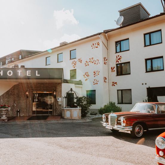 Der Eingang eines modernen Hotels mit dekorativen Blättern und Fenstern, mit einem klassischen Auto vor dem Gebäude unter einem klaren Himmel.