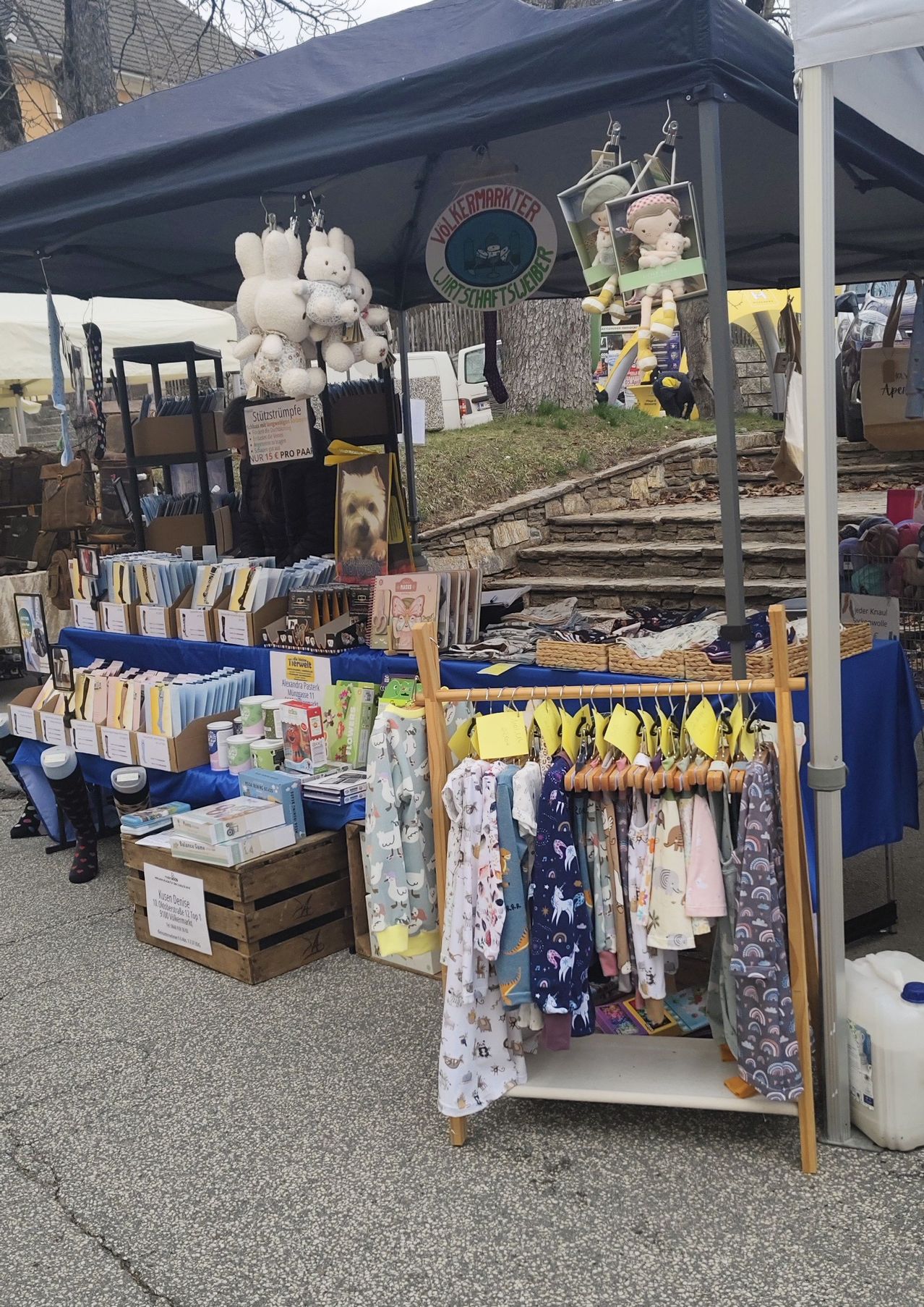 Ein Stand auf einem Markt bietet verschiedene Artikel an. Gezeigt werden Bücher, Kleidung und Kuscheltiere. Ein blauer Tischläufer bedeckt den Tisch. Ein Korb und Lagerboxen liegen auf dem Boden.