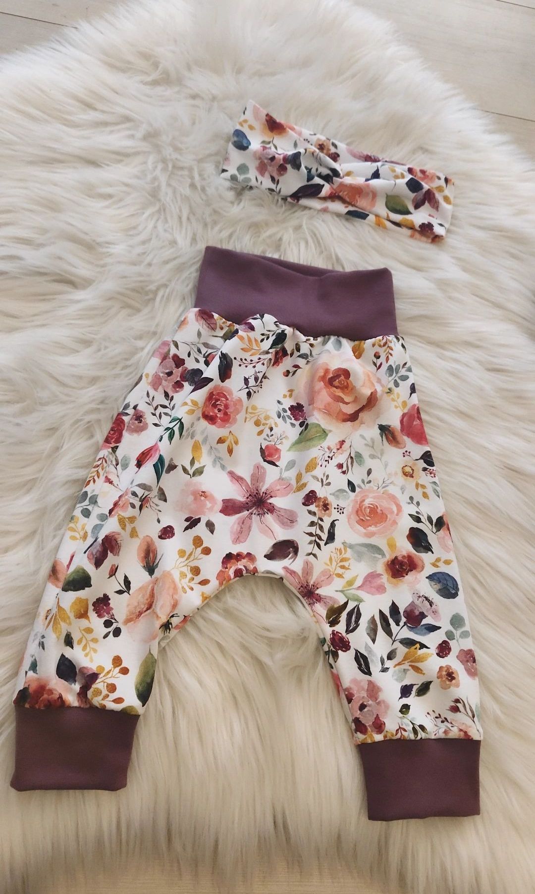 A pair of floral-printed baby pants with a purple waistband on a white furry surface, accompanied by a matching headband.