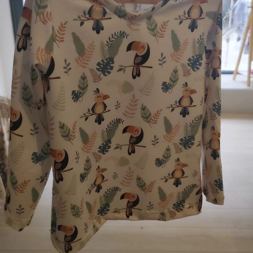 A beige long-sleeve shirt with a jungle-themed print of birds and leaves hangs on a hanger. The shirt has a relaxed fit and is made of soft fabric.