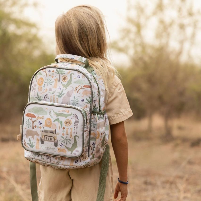 Ein junges Mädchen mit blonden Haaren geht in einem Feld, trägt ein khakifarbenes Hemd und einen Rucksack mit Safari-Tier-Print.