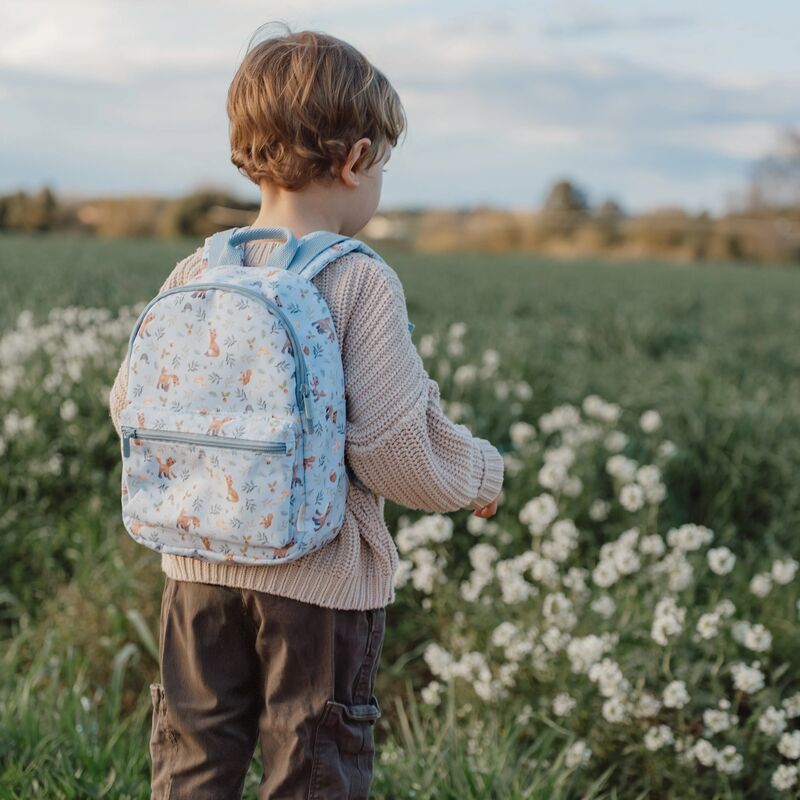 Ein junges Kind mit einem Rucksack steht in einem Feld weißer Blumen. Das Kind trägt einen hellen Pullover und braune Hosen.