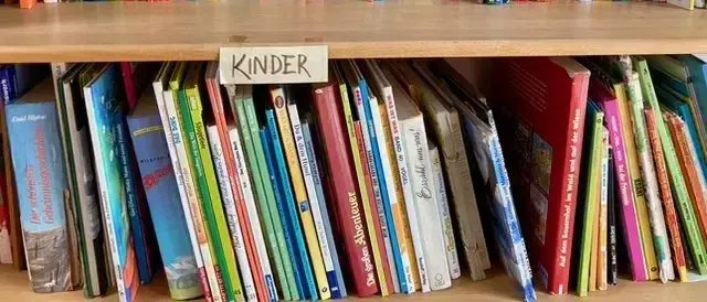 Ein holzfarbenes Regal mit zwei Ebenen zeigt Bücher. Das obere Regal hat ein Schild, das Jugend steht. Das untere Regal hat ein Schild, das Kinder steht. Die Bücher sind in Reihen angeordnet.