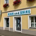Ein gelbes Gebäude mit einem Schild 'Nimm und Bring', zwei Fenstern, einer Tür und Blumenkästen mit roten Blumen.
