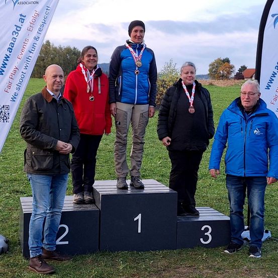 Fünf Personen stehen auf einem Podest mit den Nummern 1, 2 und 3. Sie tragen Medaillen und lächeln. Der Mann links trägt einen schwarzen Mantel, und der Mann rechts einen blauen Mantel. Dahinter stehen zwei Banner mit Logos.