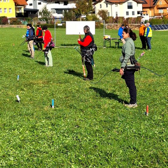 Eine Gruppe von Menschen befindet sich auf einem Rasenfeld, möglicherweise zum Bogenschießen. Einige halten Bögen und Pfeile, während andere zusehen. Ein Tisch mit Gegenständen und eine weiße Tafel sind in der Nähe.