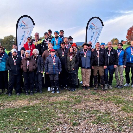 Eine Gruppe von Bogenschützen mit Medaillen steht auf einem Rasenfeld vor einem Hintergrund mit bewölktem Himmel. Zwei Banner mit Logos und Texten stehen hinter ihnen.