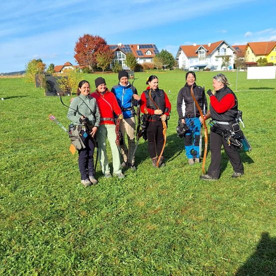 Sechs Frauen stehen auf einem Rasenfeld mit Bögen und Pfeilen, lächeln und tragen Winterjacken. Hinter ihnen stehen Häuser, Bäume und ein weißes Schild.