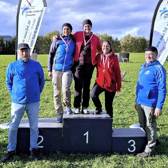 Fünf Personen stehen auf einem Podest mit Medaillen, zwei stehen oben. Die Frau links trägt eine blaue Jacke und beige Hose. Die Frau rechts trägt eine rote Jacke. Ein Mann steht links und einer rechts.