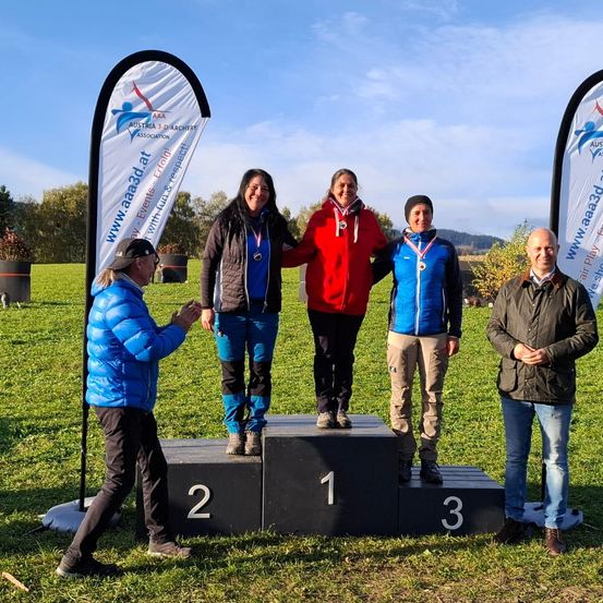 Fünf Personen stehen auf einem Podest mit Medaillen, zwei Bannern dahinter und einem Rasenfeld im Hintergrund.